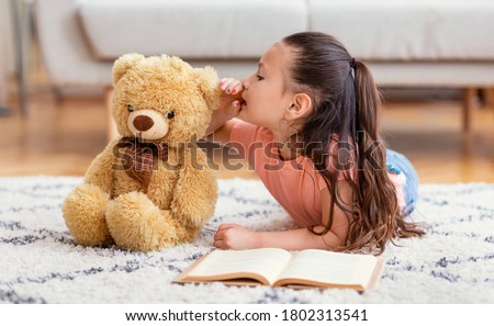 Asian Kid Girl Sharing Secret With Teddy Bear Toy Playing Reading Book Lying On Floor At Home. Best Friend Concept