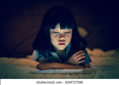 Asian Kid Girl Using Tablet Watching Movie While Lying On Bed, In Dark Background Of Bedroom, Bright Light Screen Reflex On Her Face.