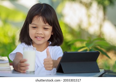 Asian Kid Girl,age 4 To 6 Years Old, Looks Cute, Bright And Strong.In His Hand Was A Glass Of Milk. There Are Tablets And Books On The Table. She Is Studying Online And Drinking Milk.Green Background 