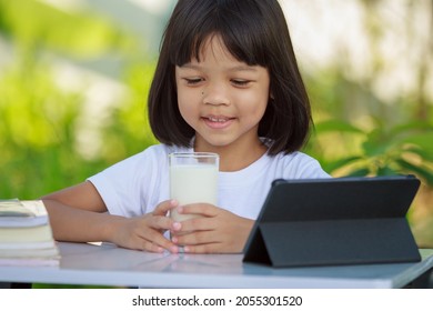 Asian Kid Girl,age 4 To 6 Years Old, Looks Cute, Bright And Strong.In His Hand Was A Glass Of Milk. There Are Tablets And Books On The Table. She Is Studying Online And Drinking Milk.Green Background 