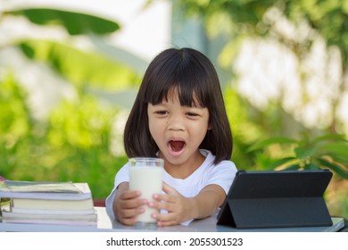 Asian Kid Girl,age 4 To 6 Years Old, Looks Cute, Bright And Strong.In His Hand Was A Glass Of Milk. There Are Tablets And Books On The Table. She Is Studying Online And Drinking Milk.Green Background 