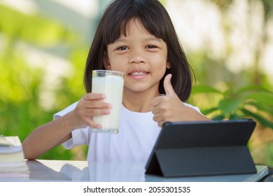 Asian Kid Girl,age 4 To 6 Years Old, Looks Cute, Bright And Strong.In His Hand Was A Glass Of Milk. There Are Tablets And Books On The Table. She Is Studying Online And Drinking Milk.Green Background 