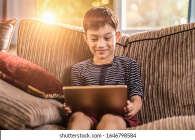 Asian Kid Holding And Playing On His Ipad Tablet Device Sitting On Sofa With Pillow, Smiling Joyfully Enjoying Free Time, Sunset Jour With Light Shining In Living Room, Wearing Short And Stripy Shirt