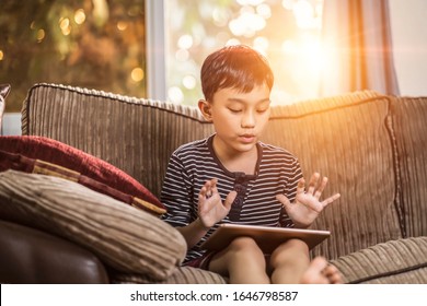 Asian Kid Holding And Playing On His Ipad Tablet Device Sitting On Sofa With Pillow, Smiling Joyfully Enjoying Free Time, Sunset Jour With Light Shining In Living Room, Wearing Short And Stripy Shirt