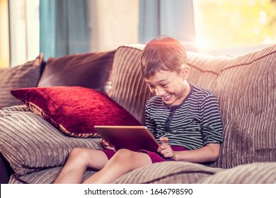 Asian Kid Holding And Playing On His Ipad Tablet Device Sitting On Sofa With Pillow, Smiling Joyfully Enjoying Free Time, Sunset Jour With Light Shining In Living Room, Wearing Short And Stripy Shirt