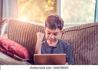 Asian Kid Holding And Playing On His Ipad Tablet Device Sitting On Sofa With Pillow, Smiling Joyfully Enjoying Free Time, Sunset Jour With Light Shining In Living Room, Wearing Short And Stripy Shirt