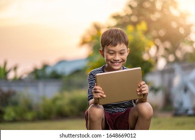 asian kid holding playing smart tablet device playing games, sitting outdoor in garden, smiling joyfully enjoying free time, sunset shining through bokeh background, wearing short and stripy shirt - Powered by Shutterstock