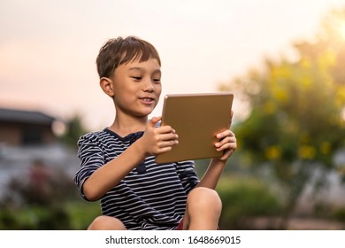 asian kid holding playing smart tablet device playing games, sitting outdoor in garden, smiling joyfully enjoying free time, sunset shining through bokeh background, wearing short and stripy shirt - Powered by Shutterstock