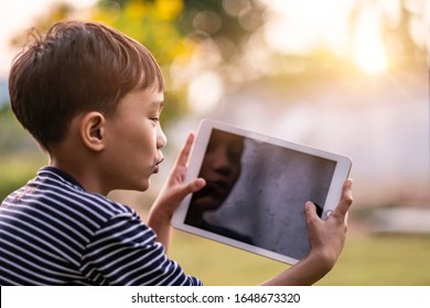 asian kid holding playing smart tablet device playing games, sitting outdoor in garden, smiling joyfully enjoying free time, sunset shining through bokeh background, wearing short and stripy shirt - Powered by Shutterstock