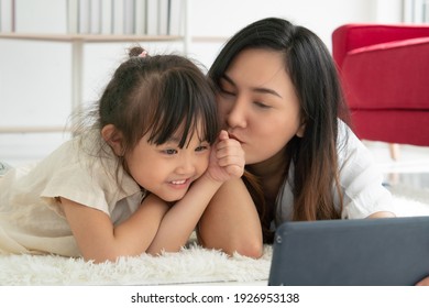 Asian Kid Kissing Her Mom While The Mother Using Tablet To Taking Photo Of Them In Living Room. Family Together Concept