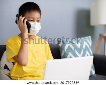 asian kid studying remotely in living room at home with a laptop. The boy is talking on the phone with the coach and typing on a laptop