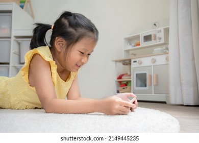 Asian Kid Using Mobile Phone On A White Carpet. School Girl Laying Down On The Floor Happily Using Social Network Or Playing Game.