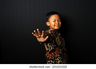 Asian Kid Wearing Batik Waving To The Camera Isolated On Black Background.