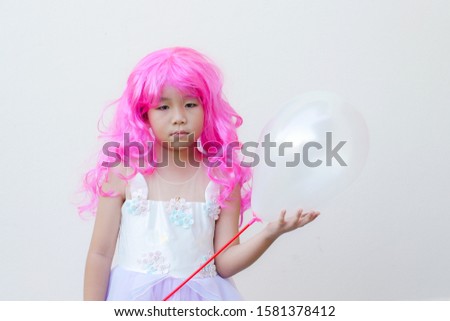 Asian kid wears a pink wig,a fancy dress and hold a white balloon.
