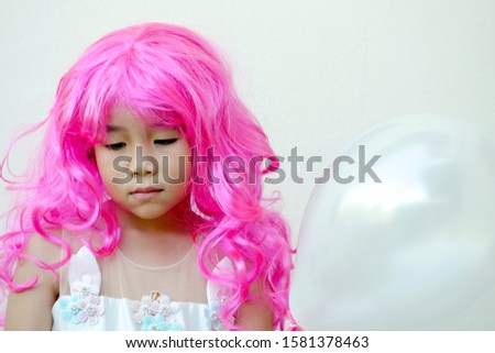 Asian kid wears a pink wig,a fancy dress and hold a white balloon.