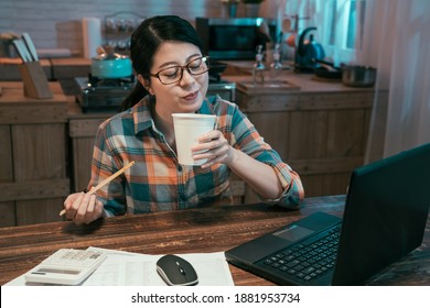 Asian Korean Businesswoman Eating Instant Noodles At Kitchen For Bedtime Snack Time. Charming Female Enjoy Ramen Soup In Paper Cup Smiling. Happy Lady With Unhealthy Food And Laptop Working In Night