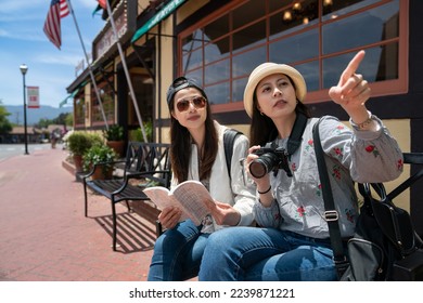 asian korean woman holding camera showing direction with pointing gesture as her travel companion is using guidebook on public bench in solvang. they are discussing next destination to go - Shutterstock ID 2239871221