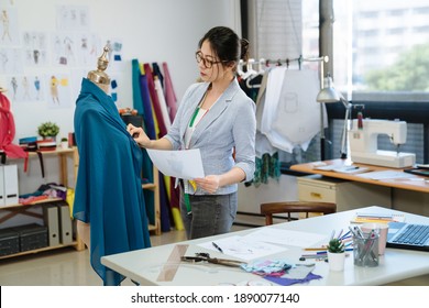 asian korean woman tailor in seamstress workshop working. stylish female fashion designer with measuring tape on neck looking at mannequin and sketch paper in studio. girl staff checking with draw - Powered by Shutterstock