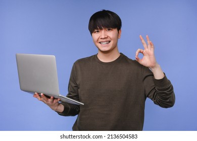 Asian korean young coder programmer student man freelancer hacker working remotely on laptop, doing online shopping, e-learning on distance, browsing showing okay isolated in blue background