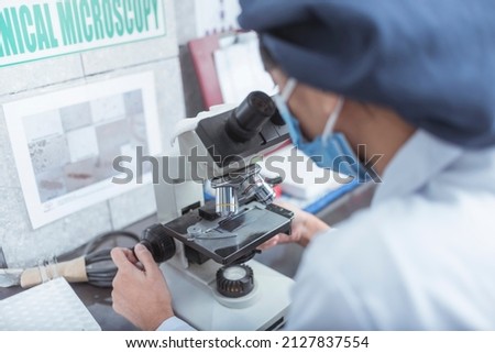 An asian laboratory technician checks a swab specimen sample with an optical microscope. Working at a research facility of hospital lab. Wearing a bouffant cap and face mask.