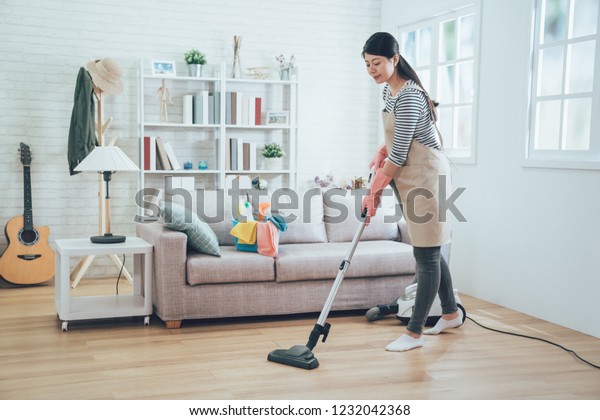 asian lady doing house chores in apron. young\
housewife using vacuum cleaner cleaning the wooden floor in the\
living room. happy housekeeper doing housework at home with\
attractive smile on face.