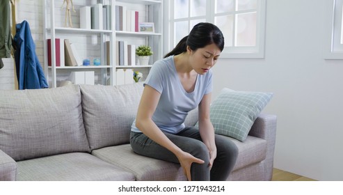 Asian Lady Frowning Massage Hurts Knee In Sofa In Living Room In Apartment. Young Woman Suffering From Leg Pain With Painful Face. Beautiful Housewife Carefully Touching Her Knees By Hands.