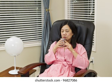 Asian Lady Holding Cup Of Coffee In Bathrobe While Relaxing In Massage Chair At Home