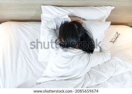 Asian Lady in white long arm t-shirt pajamas lie prone, lie on the stomach in bed with tried and exhausted mood and tone.