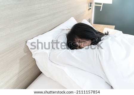 Asian Lady in white long arm t-shirt pajamas lie prone, lie on the stomach in bed with tried and exhausted mood and tone.