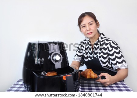 an asian lady woman is cooking her croissant by oil - free air fryer machine for breakfast in the kitchen with white cement wall background