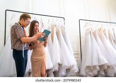  Asian LGBTQ Guy Bridal Shop Owner Using Digital Tablet Helping Female Customer Choosing Wedding Gown At Wedding Studio. Small Business Entrepreneur Wedding Planner And Marriage Ceremony Concept