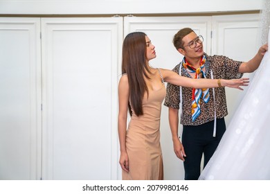 Asian LGBTQ Guy Bridal Shop Owner Helping Female Bride Customer Choosing And Trying On Wedding Gown At Wedding Studio. Small Business Entrepreneur Wedding Planner And Marriage Ceremony Concept