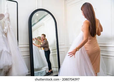 Asian LGBTQ Guy Bridal Shop Owner Helping Female Bride Customer Choosing And Trying On Wedding Gown At Wedding Studio. Small Business Entrepreneur Wedding Planner And Marriage Ceremony Concept