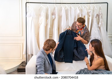 Asian LGBTQ Guy Bridal Shop Owner Using Laptop Help Male And Female Couple Customer Choosing Wedding Gown And Suit At Wedding Studio. Small Business Entrepreneur Wedding Planner And Marriage Concept