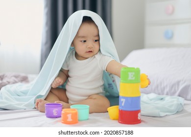 Asian Little Baby Boy Playing A Tower Cup Toy On A Bed In Bedroom. A Kid Playing And Smiling Under Covering A Blue Duvet. Child Development And Creativity Concept.