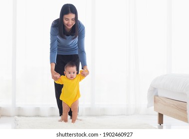 Asian Little Baby Girl Taking First Steps Learning To Walk With Mom Help Support The Cute Unstable Walking Toddler At Home In Bedroom. Happy Family First Steps Parenthood Concept