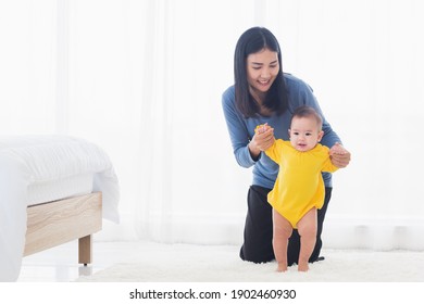 Asian Little Baby Girl Taking First Steps Learning To Walk With Mom Help Support The Cute Unstable Walking Toddler At Home In Bedroom. Happy Family First Steps Parenthood Concept