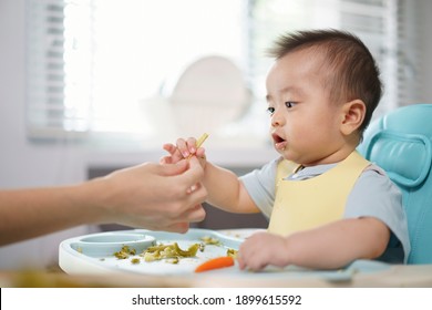 Asian Little Baby Receive A Baby Corn From Her Hand Mother. A Son Seat On A High Chair In The Kitchen At Home. A Boy Looking At Her Hand Mom Holding A Baby Corn.