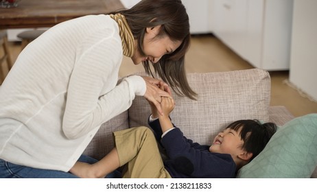 Asian Little Boy Laughing And Trying To Pushing His Mother Away While She Is Playing Tickle With Him On The Couch In The Living Room At Home