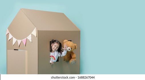 Asian little child girl playing with cardboard house with her teddy bear  isolated on blue long banner with copy space for your text, Creative at home with family concept