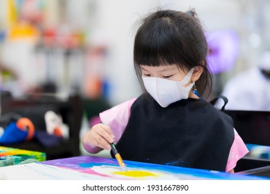 Asian Little Child Girl Wearing A 3D Surgical Face Mask. Children Learn Art And Paint On Canvas. Kid Wears A Black Apron. Happy Study In A Combined Class. Pupil Aged 4 Years Old.