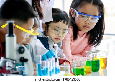 Asian Little Curious Preschool Girl Focusing Observing At Teacher Pouring Red Reagent From Glass Beaker Do Chemical Experiment On Laboratory Table With Microscope And Equipment With Young Friends.