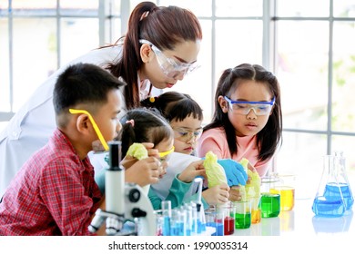 Asian Little Curious Preschool Girl Focusing Observing At Teacher Pouring Red Reagent From Glass Beaker Do Chemical Experiment On Laboratory Table With Microscope And Equipment With Young Friends.