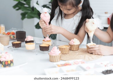 Asian little cute child girl enjoy making cupcakes with friend at home kitchen, squeezing cream from piping bag to decorate homemade cupcake, smiling with holiday activities. Selective focus on hands - Powered by Shutterstock