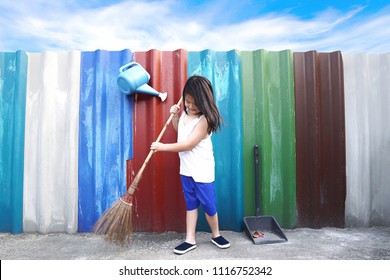 Asian Little Cute Girl Sweeping The Floor With Broom At Outdoor On Colourful Wall And Blue Sky Background.Inspired Kid Doing Household Chore.People,Housework, Cleaning And Housekeeping Concept. 