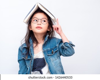 Asian Little Cute Girl Wearing Glasses And Put The Book On Head. Preschool Lovely Kid With The Book Covering On Her Head And Pointing Finger Up. Learning And Education Of Kid.