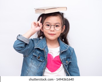 Asian Little Cute Girl Wearing Glasses And Put The Book On Head. Preschool Lovely Kid With The Book On Her Head And Pointing Finger Up. Learning And Education Of Kid.