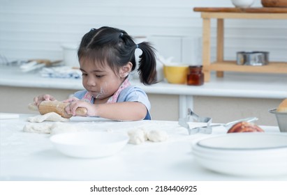 Asian Little Cute Kid Girl Wear Apron With Flour Mess Up On Face And Arms Have Fun Holding Wooden Rolling Pin Kneading Dough Bread In Kitchen At Home Or Baking Class At School. Child Education Concept