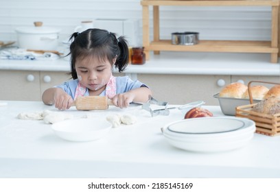 Asian Little Cute Kid Girl Wear Apron With Flour Mess Up On Face And Arms Have Fun Holding Wooden Rolling Pin Kneading Dough Bread In Kitchen At Home Or Baking Class At School. Child Education Concept