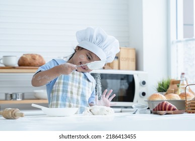 Asian Little Cute Kid Girl Wear Chef Hat And Apron With Flour Mess Up On Face Have Fun Sifting Flour On Dough Bread In Kitchen At Home Or Baking Class At School. Child Education Concept